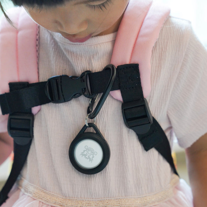 A young child wearing a pale pink dress carries a pink backpack with black straps and a plastic buckle, featuring the Catalyst Case US AirTag - Total Protection Case, Hang-It.