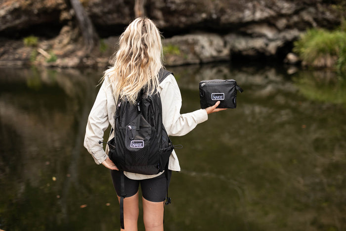 Black foldable 30L backpack with white NAKIE logo, shown worn by person holding matching black pouch by water.
