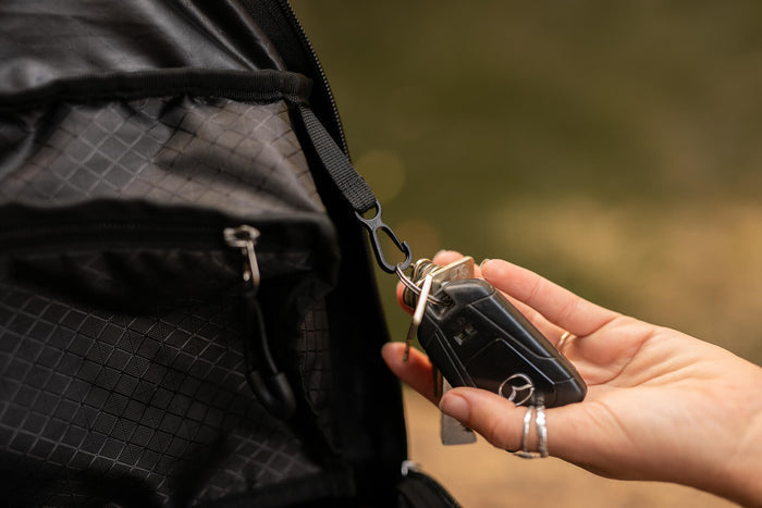 Black foldable backpack with quilted texture and attached black webbing strap and keychain clip, shown being held