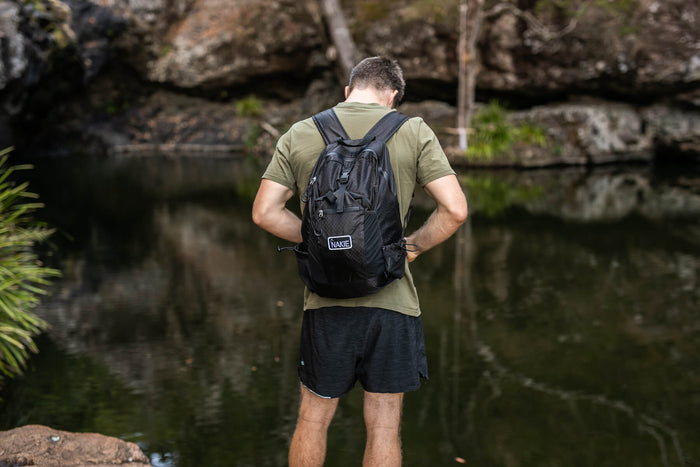 Black foldable backpack with branded patch, worn on back by person at water's edge