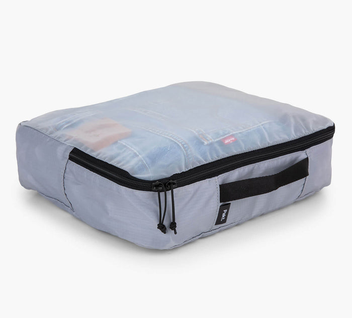 Three-quarter view of grey packing cube with translucent top showing folded clothing, black zipper and side handle on white background