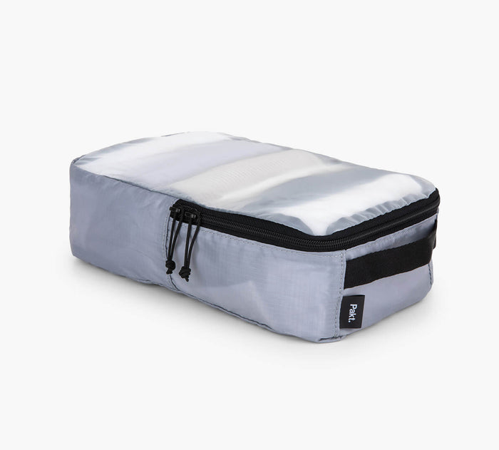 Three-quarter view of grey packing cube showing translucent top panel, black zipper, and side handle on white background