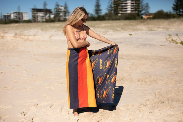 Adelaide Crows AFL recycled beach towel with navy, orange, and gold stripe design, displayed on sand.