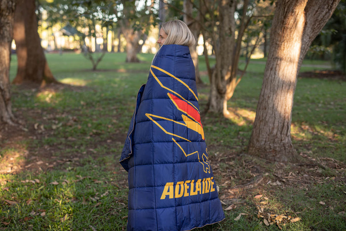 Navy puffy down blanket with Adelaide Crows logo and red-gold lightning bolt design, shown wrapped around person from behind.
