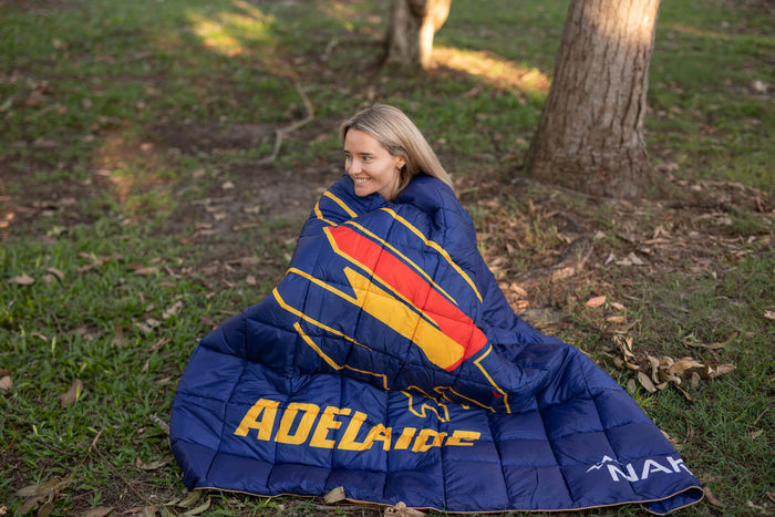 Navy puffy blanket with Adelaide Crows AFL design, red and gold stripe pattern, displayed outdoors wrapped around woman.