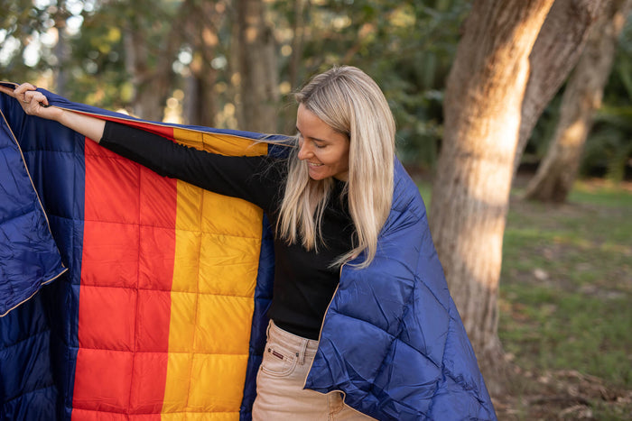 Navy quilted down puffy blanket with black, red, and gold striped pattern, displayed outdoors wrapped around a woman.