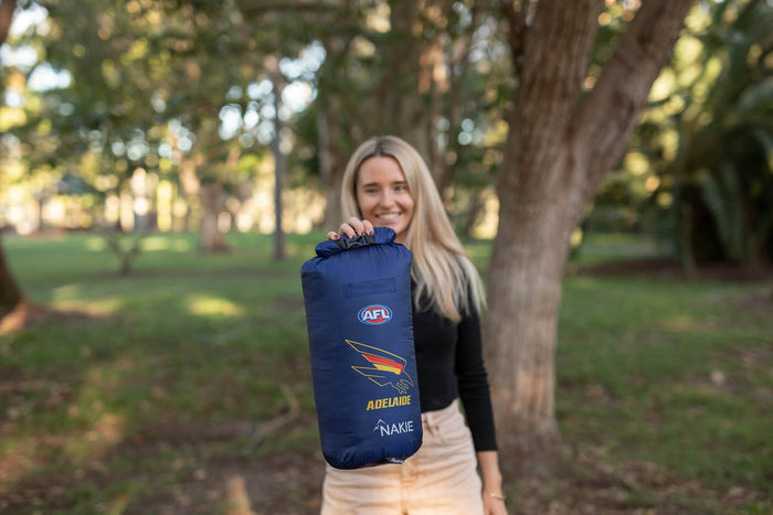 Navy down puffy blanket with AFL Adelaide Crows logo and Nakie branding, shown rolled and held up outdoors.