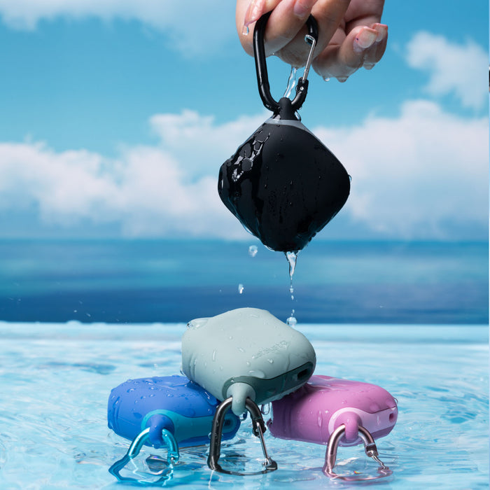 Near the pool, four vibrant waterproof devices in blue, green & pink AirPods 5/4 waterproof cases with a smooth one-piece design and handy carabiners are displayed. A hand holds a Catalyst AirPods 5/4 in a black waterproof case, its silicone exterior gleaming with water droplets to highlight its durability and style.