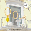 This image shows a balcony background with a set of three earrings. This set contains a lemon charm huggie, topaz pave clicker, and micro topaz flatback stud. All of these earrings are 14k gold plated.