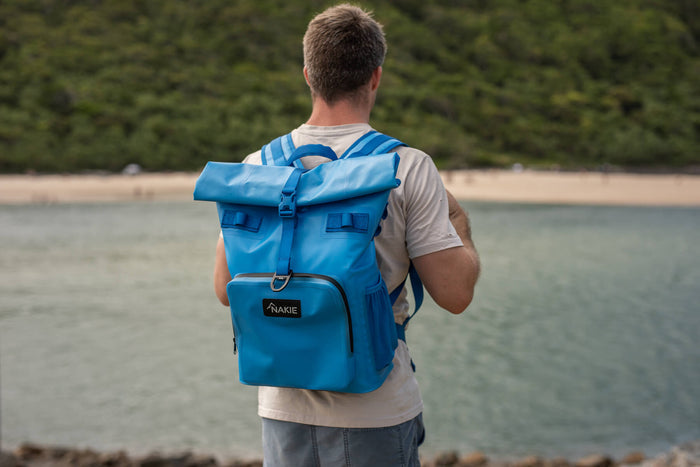 Blue cooler backpack with roll-top closure worn by man at beach, featuring front pocket and shoulder straps.
