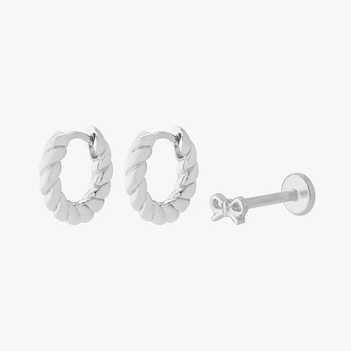 This image shows a set of silver earrings.  The set includes a pair of silver french twist huggies and a singular bow flatback stud.