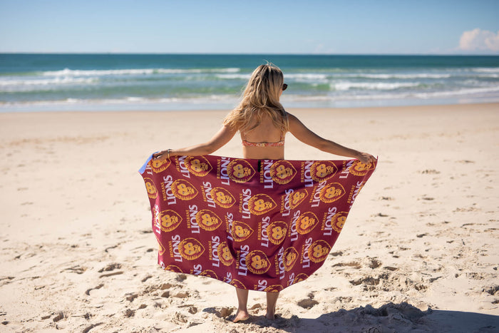 Burgundy beach towel with Brisbane Lions AFL logo and lion head repeat pattern, draped on person's shoulders at beach