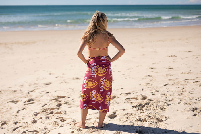Burgundy beach towel with Brisbane Lions AFL logo and text pattern, worn as a wrap on person standing on sand facing ocean