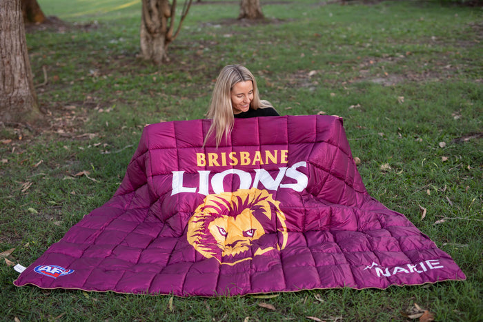 Maroon quilted puffy blanket with Brisbane Lions logo, text, and lion head graphic displayed outdoors on grass.