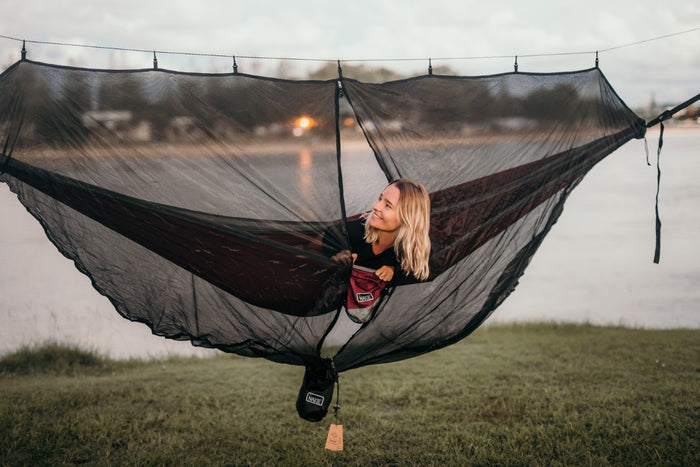 A smiling woman peeking out from inside Nakie's Bug Net while lying in a hammock