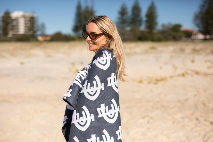 Carlton Blues recycled sand-free beach towel in charcoal with white geometric logo pattern, worn as a wrap on beach