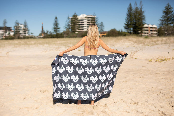 Navy beach towel with white repeating logo pattern, held open on sand with beachfront buildings in background