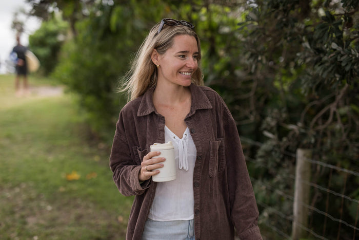 Cream coffee cup lid on reusable cup held by woman wearing brown corduroy shirt outdoors