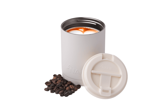 White insulated coffee cup with latte art, cream lid removed, and coffee beans scattered beside it