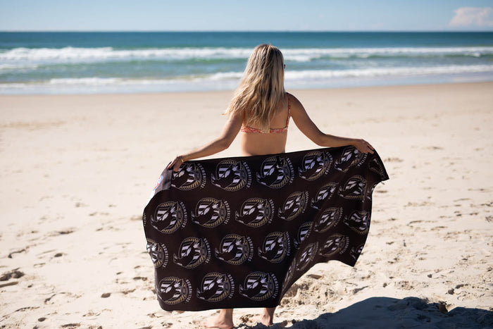 Black Collingwood Magpies AFL beach towel with circular logo pattern, displayed on sandy beach