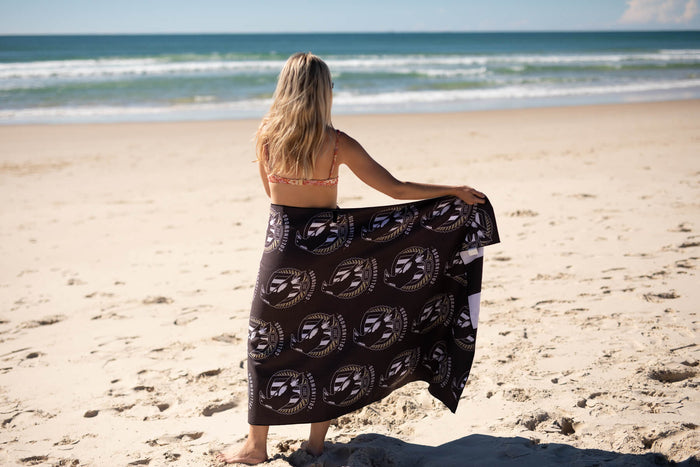 Black beach towel with Collingwood Magpies AFL circular logo repeat pattern, shown draped on person at beach