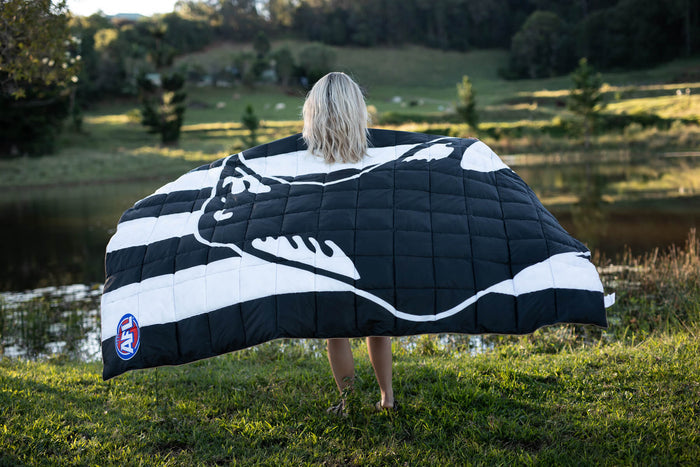 Black and white striped quilted blanket with Collingwood Magpies magpie logo, worn as cape outdoors by water.