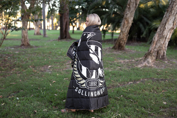 Black quilted puffy blanket with Collingwood Magpies logo and white wing design, worn over shoulders outdoors.