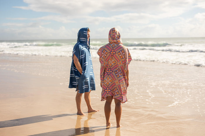 Two people wearing hooded beach towels standing in the ocean, one navy with white stripes, one red with geometric pattern.