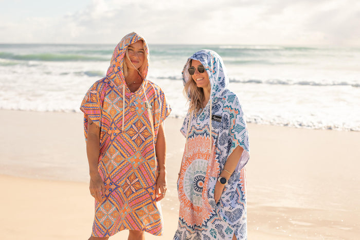 Two women wearing colorful patterned hooded towels on beach, orange geometric design and blue mandala print