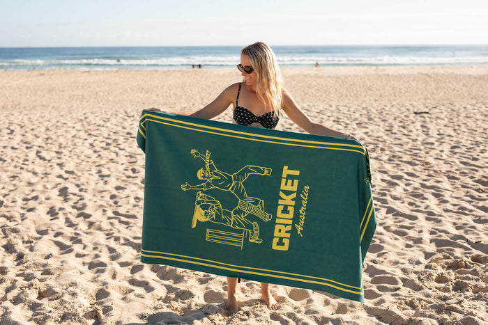 Green and gold beach towel with cricket batter graphic and Cricket Australia text, displayed on sand at the beach