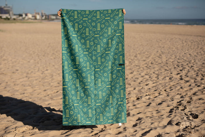 Teal recycled beach towel with cricket-themed gold pattern including bats, balls, and stumps, displayed on sand.