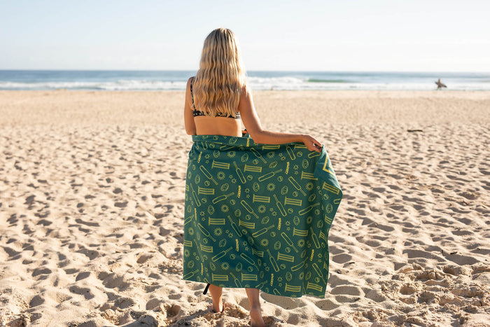 Green recycled beach towel with yellow cricket and summer pattern, shown draped on person at beach from behind.