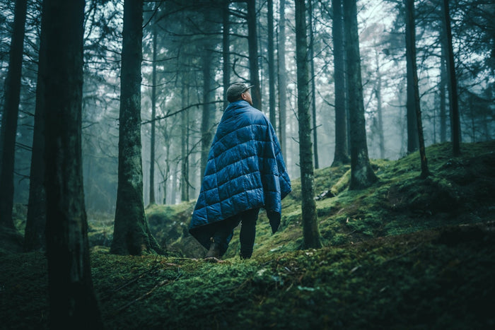 Image of a man wrapped in Nakie's Deep Ocean Blue Sustainable Down Puffy Blanket while standing in a campsite