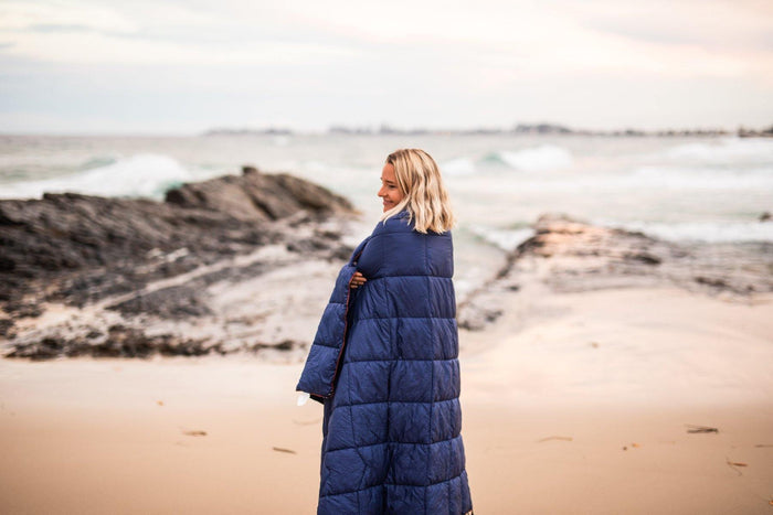 Image of a woman standing on a beach, wrapped in Nakie's Deep Ocean Blue Sustainable Down Puffy Blanket