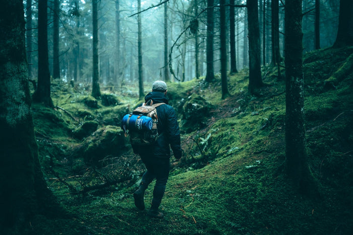 Image of a man walking in a campsite with Nakie's Deep Ocean Blue Sustainable Down Puffy Blanket attached to his backpack