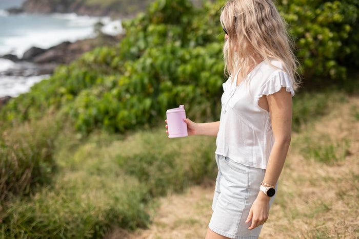 Dusty pink insulated coffee cup with lid held by woman outdoors near beach