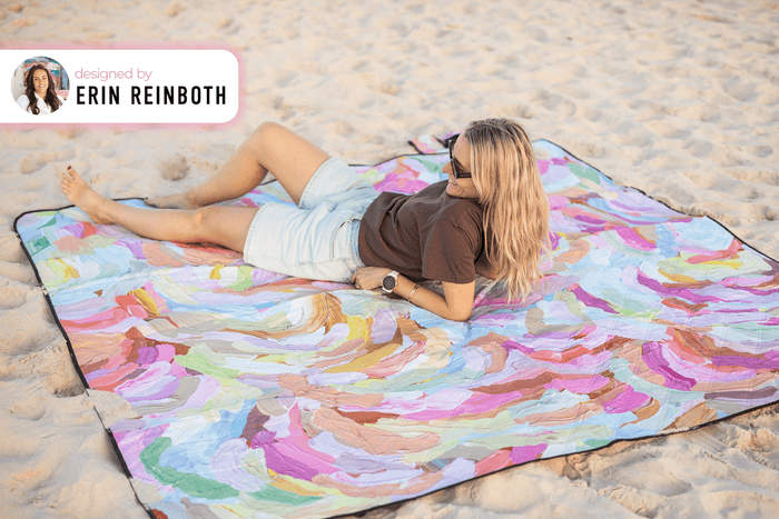 Colorful abstract painted recycled picnic blanket with multicolor brushstrokes, displayed on beach sand.