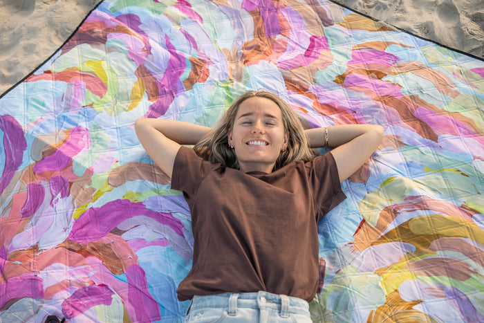 Recycled picnic blanket with colorful watercolor abstract pattern in magenta, blue, yellow, and orange tones on sand.