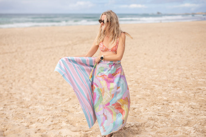 Recycled sand-free beach towel with colorful floral print, shown held on beach with striped reverse side visible.