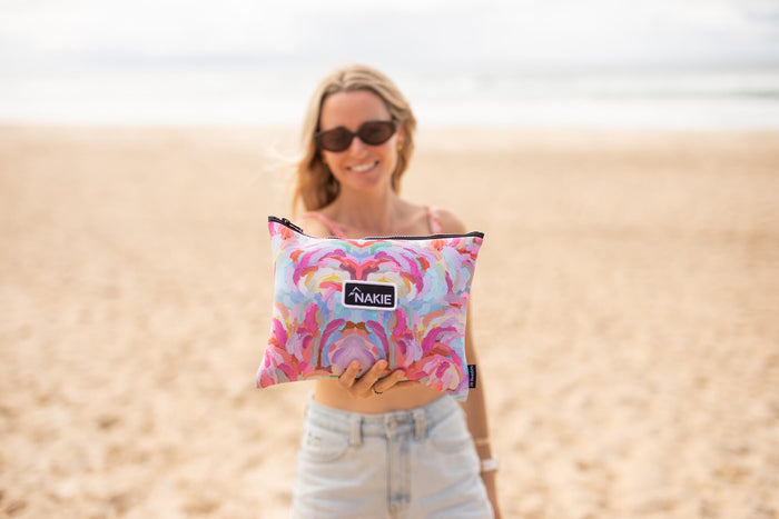 Recycled sand-free beach towel in pink and purple abstract floral print with dark trim, held by woman on beach.