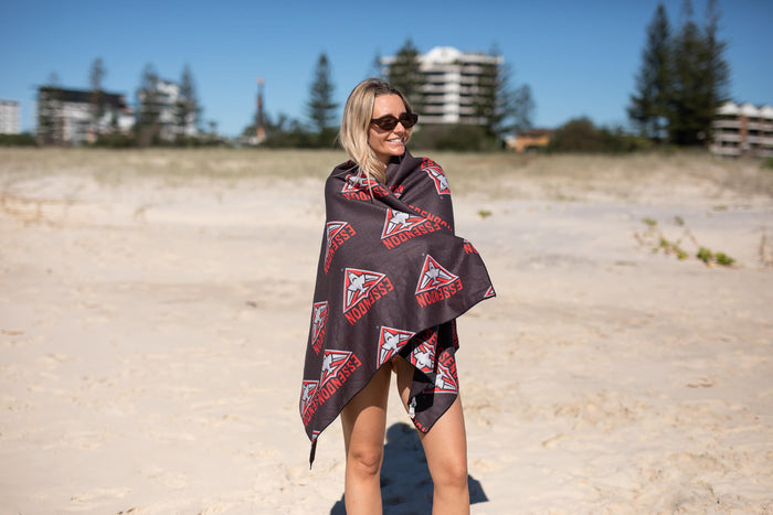 Essendon Bombers black beach towel with red and white geometric logo print, draped on woman at beach.