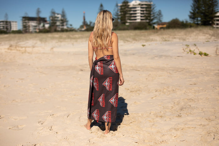 Essendon Bombers AFL patterned beach towel worn as wrap on woman at beach, black with red geometric logo design.