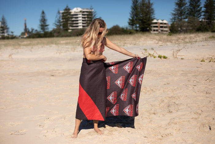 Essendon Bombers black and red beach towel with repeated triangle logo, displayed on sandy beach
