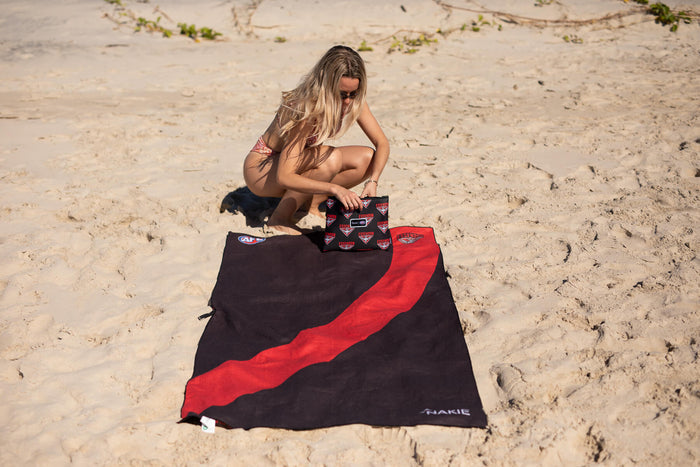 Black and red beach towel with diagonal stripe design, displayed on sand with accessories