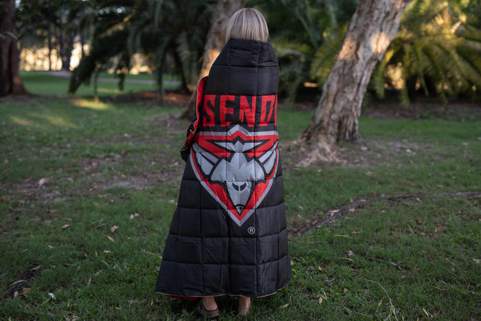 Black quilted puffy blanket with red and white Essendon Bombers logo graphic, worn as a wrap outdoors.