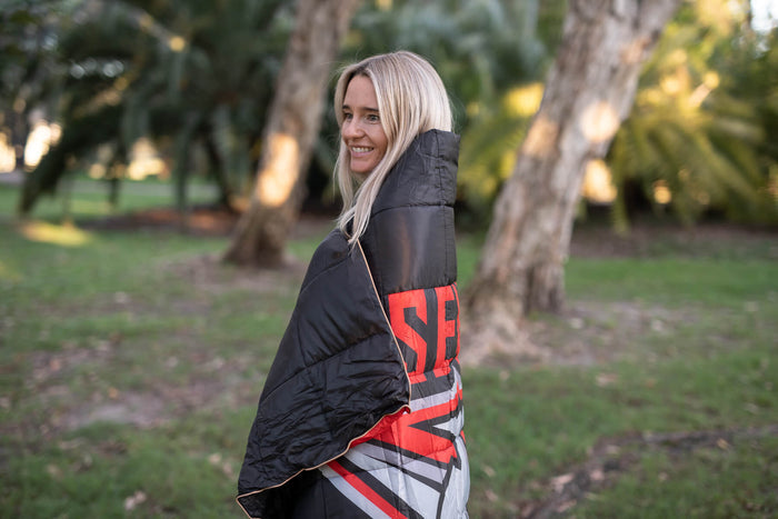 Black quilted puffy blanket with red and white Essendon Bombers AFL graphic, shown wrapped around person outdoors.