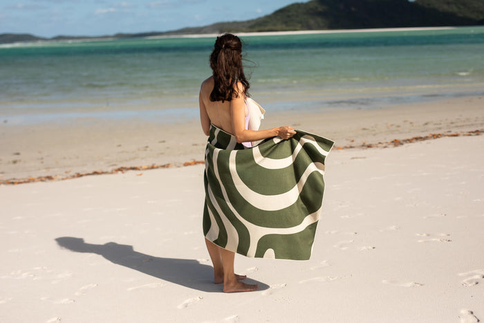Eucalyptus green beach towel with cream wavy stripes, held by woman on sandy beach overlooking turquoise water