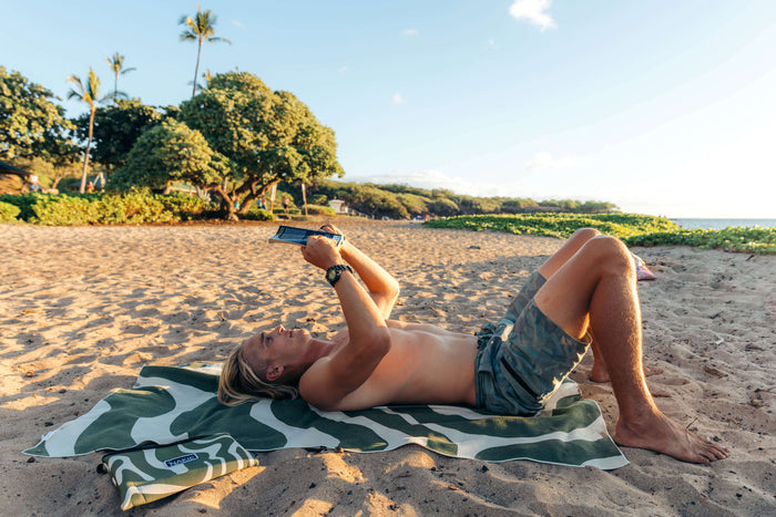 Eucalyptus green and white zebra-stripe beach towel spread on sand with person reading on it