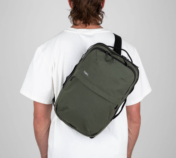 Rear three-quarter view of Everyday 15L Bag in Forest showcasing sling carry, front zip pocket, and adjustable strap on model wearing a white tee