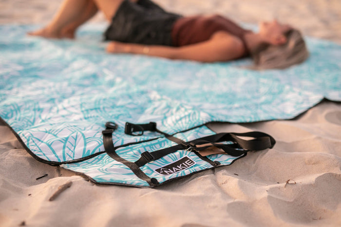 Turquoise and white leaf-patterned recycled picnic blanket with black straps, laid on sand with person in background.
